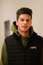 Person mit kurzgeschnittenem Haar und neutralem Gesichtsausdruck, bekleidet mit Weste.