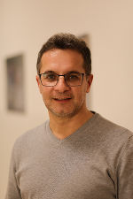 Person mit dunklem Haar und Brille trägt einen grauen Pullover. Im Hintergrund eine helle Wand.