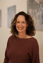 Porträt einer Person mit freundlichem Gesichtsausdruck und lockigem Haar. Trägt Schmuck und ein rotes Oberteil.