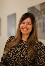 Junge Person mit langen, gewellten Haaren lächelt in die Kamera. Im Hintergrund ist ein Raum zu sehen.