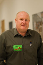 Person mit olivgrünem Hemd und Namensschild, freundlich blickend. Foto aus dem Behördenkontext.