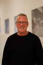 Person mit dunklem Hemd und Brille lächelt in die Kamera. Im Hintergrund ist ein Bild zu sehen.