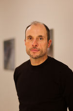 Person mit kurzem, dunklem Haar und Bart blickt in die Kamera. Im Hintergrund ein heller Raum.