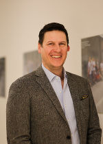 Person mit braunen Haaren in einem karierten Sakko, lächelnd. Im Hintergrund eine unscharfe Wand.