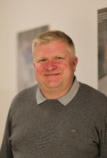 Porträt einer Person mit kurzem, hellem Haar und freundlichem Lächeln. Sie trägt einen grauen Pullover.