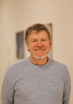 Porträt einer Person mit hellem Haar und freundlichem Lächeln. Sie trägt einen blauen Pullover.