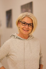 Lachende Person mit Brille und lockerem Outfit. Hintergrund ist eine helle Wand mit Kunstwerken.