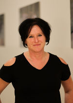 Person mit dunklen Haaren, die in die Kamera blickt. Hintergrund ist ein heller Raum mit Bildern.