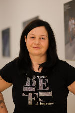 Person mit dunklen Haaren und T-Shirt schaut in die Kamera. Im Hintergrund sind Bilder zu sehen.