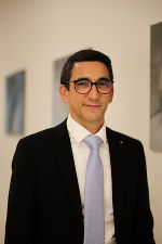 Person mit dunklem Haar und Brille in formeller Kleidung, vor einem Hintergrund mit Kunstwerken.