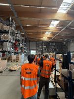 Gruppe mit Warnwesten in Lagerhalle. Hohe Regale mit Waren im Hintergrund.
