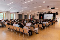 Großer Saal mit Sitzreihen, gefüllt mit Teilnehmenden einer Veranstaltung. Im Hintergrund eine Bühne mit einem Banner.